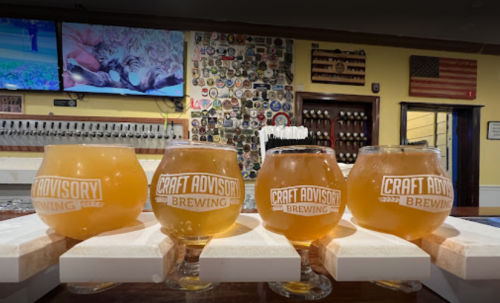 Four craft beer glasses on a bar, featuring the logo "Craft Advisory Brewing," with a wall of beer taps in the background.