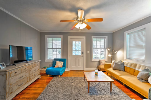 Cozy living room with a blue chair, yellow sofa, wooden coffee table, TV, and a ceiling fan, featuring natural light.