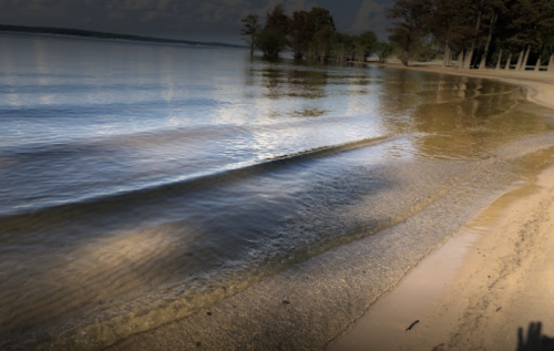 A serene lakeshore with gentle waves lapping at the sandy beach, surrounded by trees and a cloudy sky.