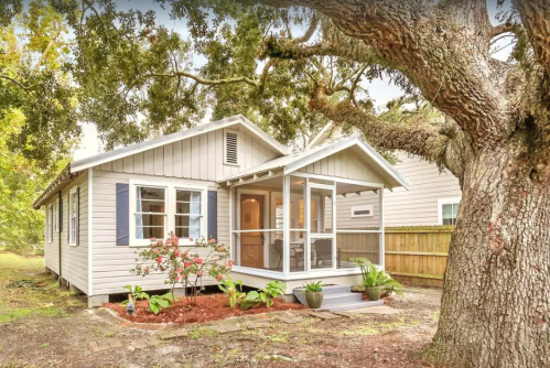 A charming single-story house with a porch, surrounded by greenery and a large tree in the front yard.