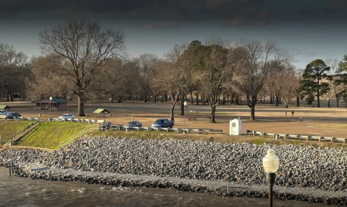 A riverside park with bare trees, a grassy area, and parked cars along a rocky shoreline under a cloudy sky.
