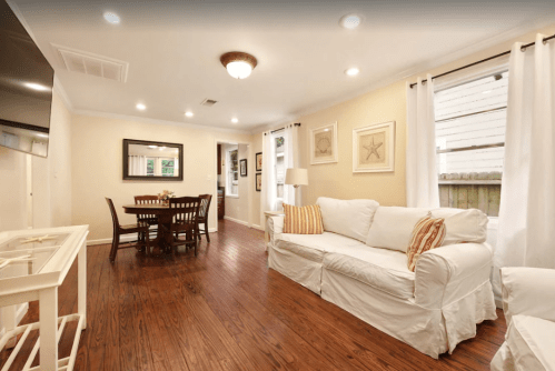 Cozy living room with a white sofa, wooden dining table, and light-colored walls, featuring natural light and decorative art.