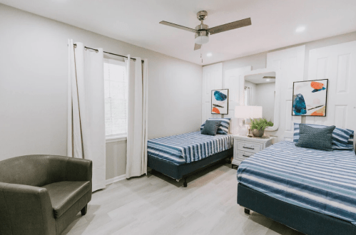 A bright bedroom with two twin beds, a chair, and modern decor, featuring light walls and natural light from a window.