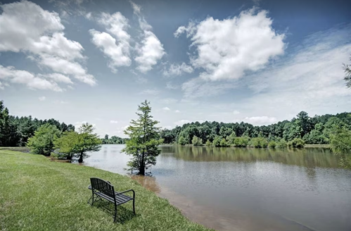 A serene lake scene with a bench by the water, surrounded by trees and a cloudy blue sky.