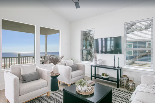 Bright, modern living room with ocean views, featuring white sofas, a black coffee table, and large windows.