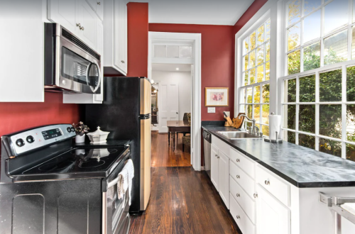 A modern kitchen with red walls, black appliances, and white cabinets, featuring a window with natural light.