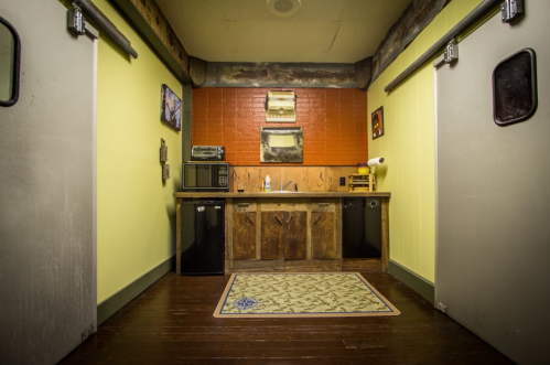 A rustic kitchen area with a wooden counter, microwave, mini fridge, and yellow walls, featuring a patterned rug.