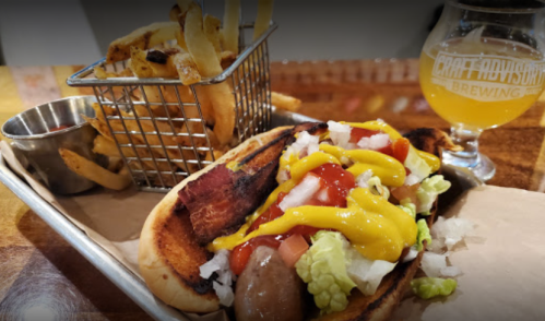 A hot dog topped with mustard, ketchup, onions, and lettuce, served with fries and a glass of beer.