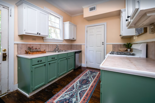 A bright kitchen with green cabinets, a patterned rug, and light-colored walls, featuring a window and door.
