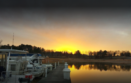 Sunset over a calm marina, with boats docked and reflections on the water, surrounded by trees and a cloudy sky.