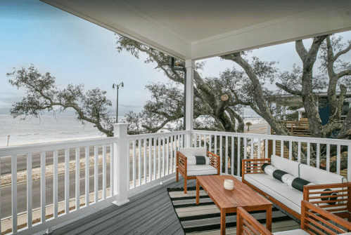 A cozy porch with wooden furniture overlooks a beach and trees, creating a serene coastal atmosphere.