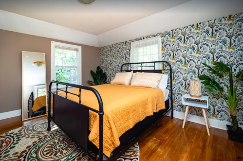 Cozy bedroom featuring a metal bed, patterned wallpaper, a mirror, and a small side table with a lamp.