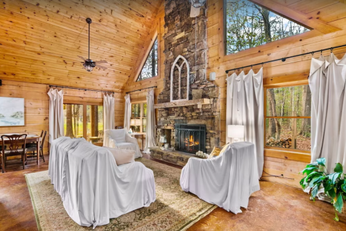 Cozy cabin interior with wooden walls, a stone fireplace, and white furniture covers, surrounded by large windows.