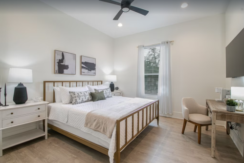Cozy bedroom with a bed, two lamps, a desk, and artwork on the walls, featuring light colors and natural light.