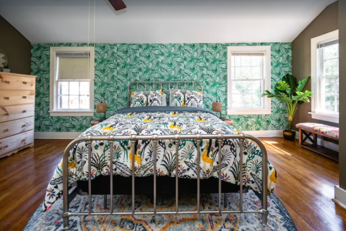 A cozy bedroom featuring a metal bed frame, vibrant bedding, green leafy wallpaper, and a potted plant by the window.