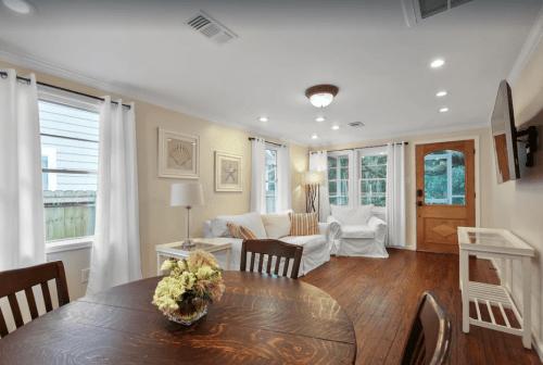 Cozy living space with a dining table, white curtains, a sofa, and a wooden door leading outside. Bright and inviting atmosphere.