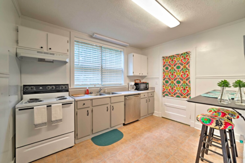 Bright kitchen with white cabinets, a stove, sink, and colorful decor, featuring a window and a small dining area.