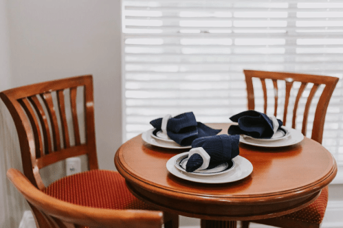 A small wooden table set for three, featuring plates and neatly folded navy napkins, with two chairs nearby.
