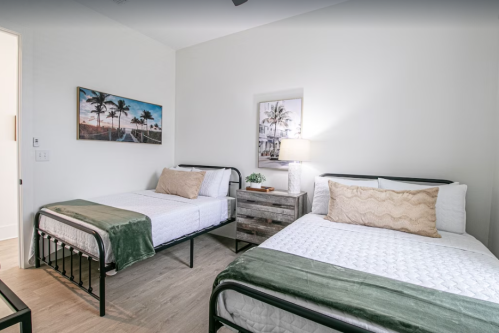 A cozy bedroom with two beds, a nightstand, and wall art featuring palm trees. Bright and inviting atmosphere.