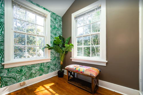 Bright corner of a room with large windows, green leaf-patterned wallpaper, and a wooden bench with colorful cushions.