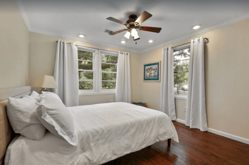 Cozy bedroom with a bed, ceiling fan, and large windows dressed in white curtains. Warm wood flooring completes the look.