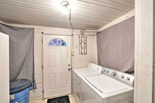 A laundry room with a washer and dryer, a door, and a blue trash can, featuring gray curtains and a light fixture.