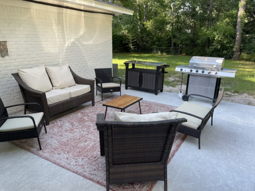 A cozy outdoor patio with seating, a coffee table, and a grill, surrounded by greenery.