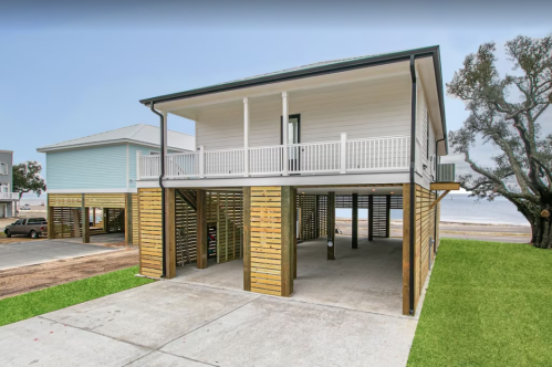 A modern beach house with a raised structure, featuring a porch and wooden accents, set near a sandy area.