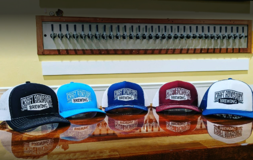 Five colorful hats with "Craft Advisory Brewing" logo displayed on a wooden counter, with beer taps in the background.