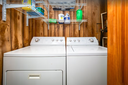 A laundry room featuring two white washing machines side by side, with shelves above holding cleaning supplies.