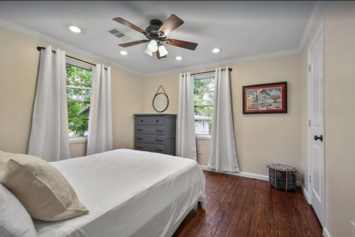 A cozy bedroom with a bed, dresser, and large windows, featuring light curtains and wooden flooring.