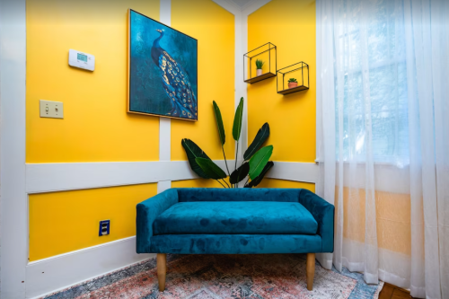 A bright yellow room features a teal couch, a peacock painting, and decorative shelves with plants.