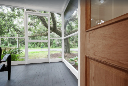 A view from a porch with a wooden door, showcasing a green landscape and trees through large windows.