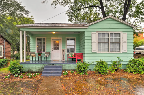 A light green house with white shutters, a red bench, and a porch surrounded by greenery on a rainy day.