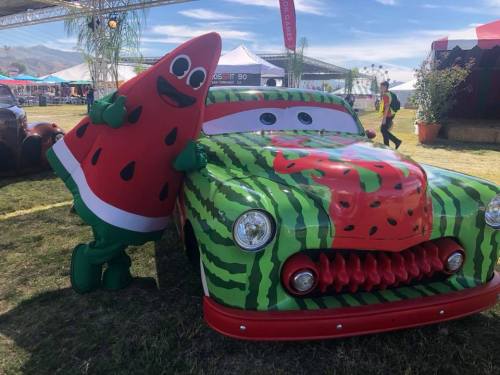 A watermelon mascot poses next to a colorful, watermelon-themed car at an outdoor event.