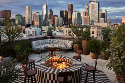 Rooftop lounge with a fire pit, surrounded by seating and city skyline views at sunset.