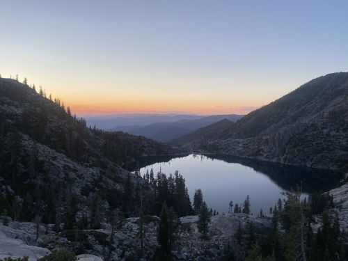 A serene mountain lake at sunset, surrounded by rocky terrain and evergreen trees, with a colorful sky in the background.