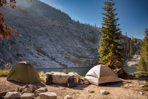Two tents set up by a serene lake, surrounded by rocky terrain and tall pine trees under a clear blue sky.