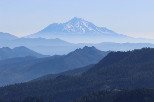 A distant snow-capped mountain rises above a layered landscape of blue hills and forests under a clear sky.