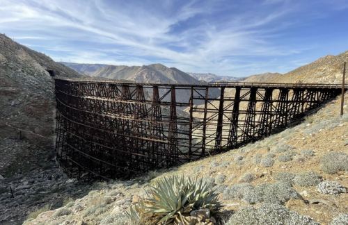 A large, weathered wooden trestle bridge spans a rocky canyon, surrounded by mountains and desert vegetation.