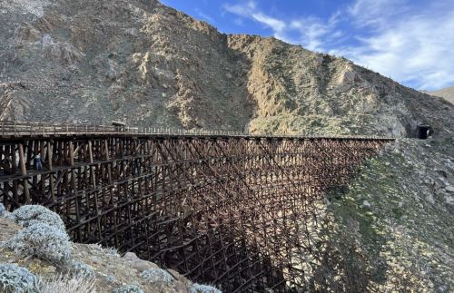 A large wooden trestle bridge spans a rocky landscape, surrounded by mountains under a blue sky with scattered clouds.