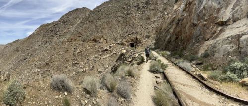 A hiker walks along a rocky trail beside old railway tracks in a mountainous desert landscape.