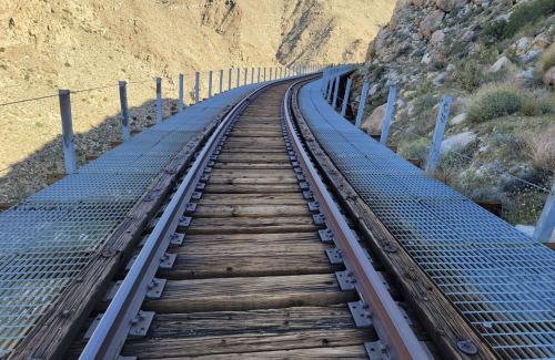 A winding railway track on a metal bridge, surrounded by rocky terrain and mountains.