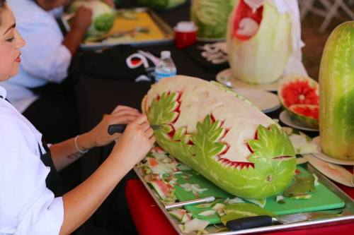 A person carves intricate designs into a large watermelon at a food festival, surrounded by other carved fruits.