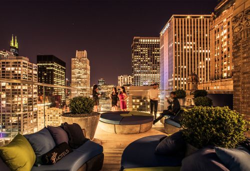 A rooftop lounge at night with city skyline views, featuring people socializing and modern seating arrangements.