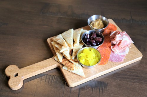 A wooden serving board with pita bread, olives, mustard, pickles, and assorted meats arranged neatly.
