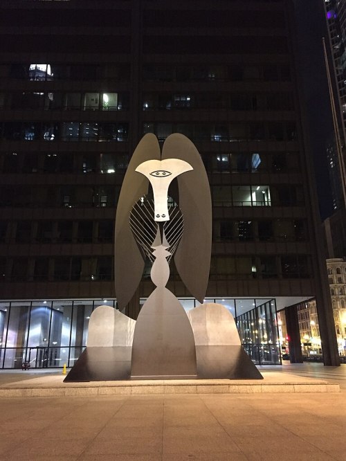 A large abstract sculpture featuring a stylized figure with wings, set against a dark urban backdrop at night.