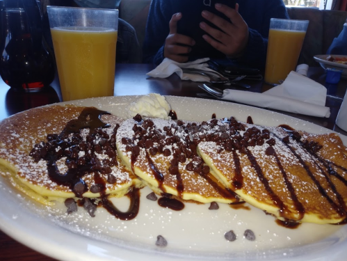 A plate of pancakes topped with chocolate chips, whipped cream, and syrup, with orange juice in the background.