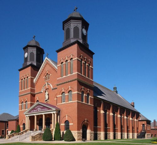 A large brick church with two clock towers and a statue above the entrance, set against a clear blue sky.