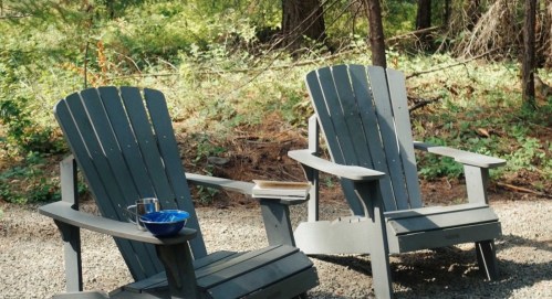 Two gray Adirondack chairs sit on a gravel path surrounded by trees, with a bowl and a book on one chair.